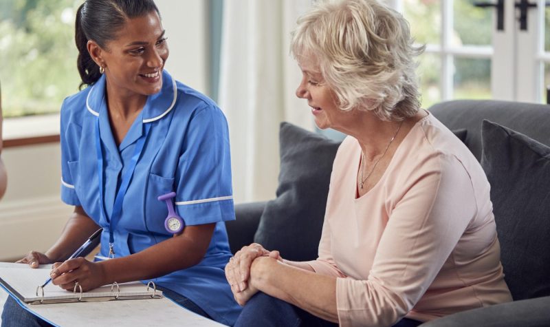 Senior Woman At Home Talking To Female Nurse Or Care Worker In Uniform Making Notes In Folder